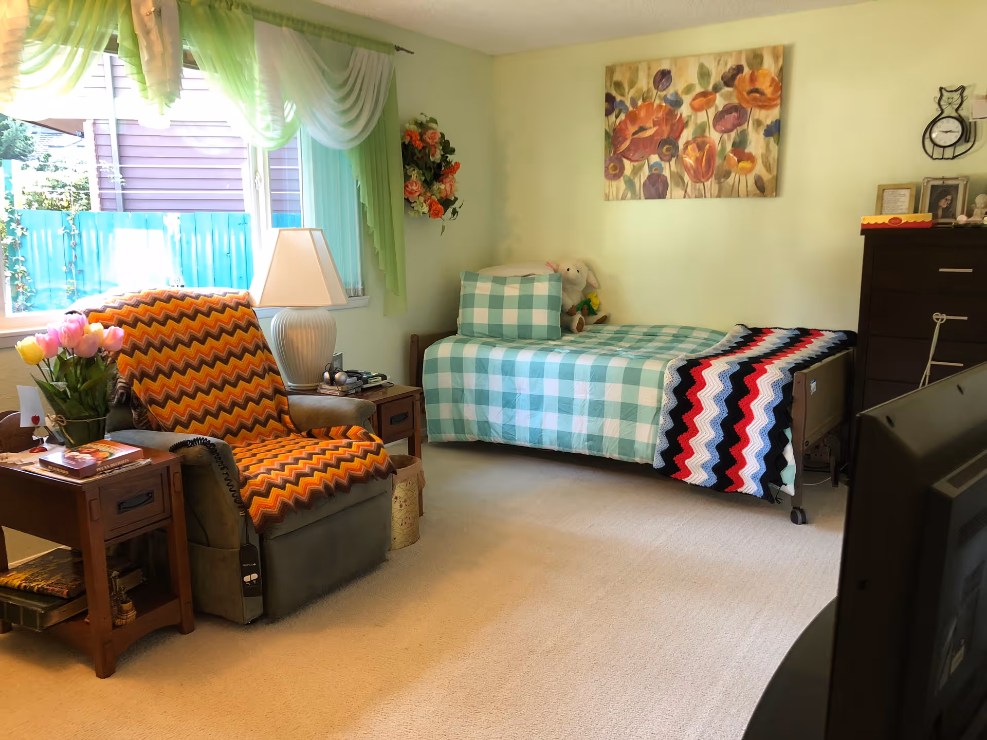 A cozy bedroom with a single bed covered in a green and white checkered bedspread and a colorful crocheted blanket. Next to the bed is a dark wooden dresser with framed photos and a clock. A comfortable armchair with an orange, yellow, and brown zigzag patterned cover sits beside a wooden side table holding a lamp, flowers, and books. The room has light green walls, a window with green and white sheer curtains, and a floral painting above the bed.