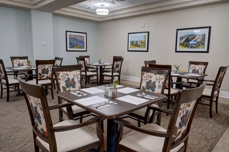A dining room with multiple square tables set with placemats, menus, silverware, and small flower vases. The chairs have wooden frames with floral patterned upholstery on the backrests. The walls are decorated with framed landscape photographs, and the room has a carpeted floor and a ceiling light fixture.
