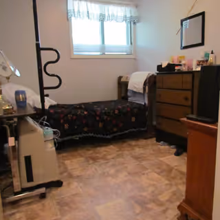 A small bedroom with a single bed covered with a dark floral blanket, a wooden dresser with various items on top, a window with white curtains, and a small TV on a wooden stand. The floor has a light brown tile pattern.
