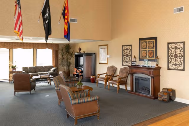 Comfortable lounge in a senior living facility with upholstered armchairs and sofas, a fireplace, decorative wall art, and flags overhead.