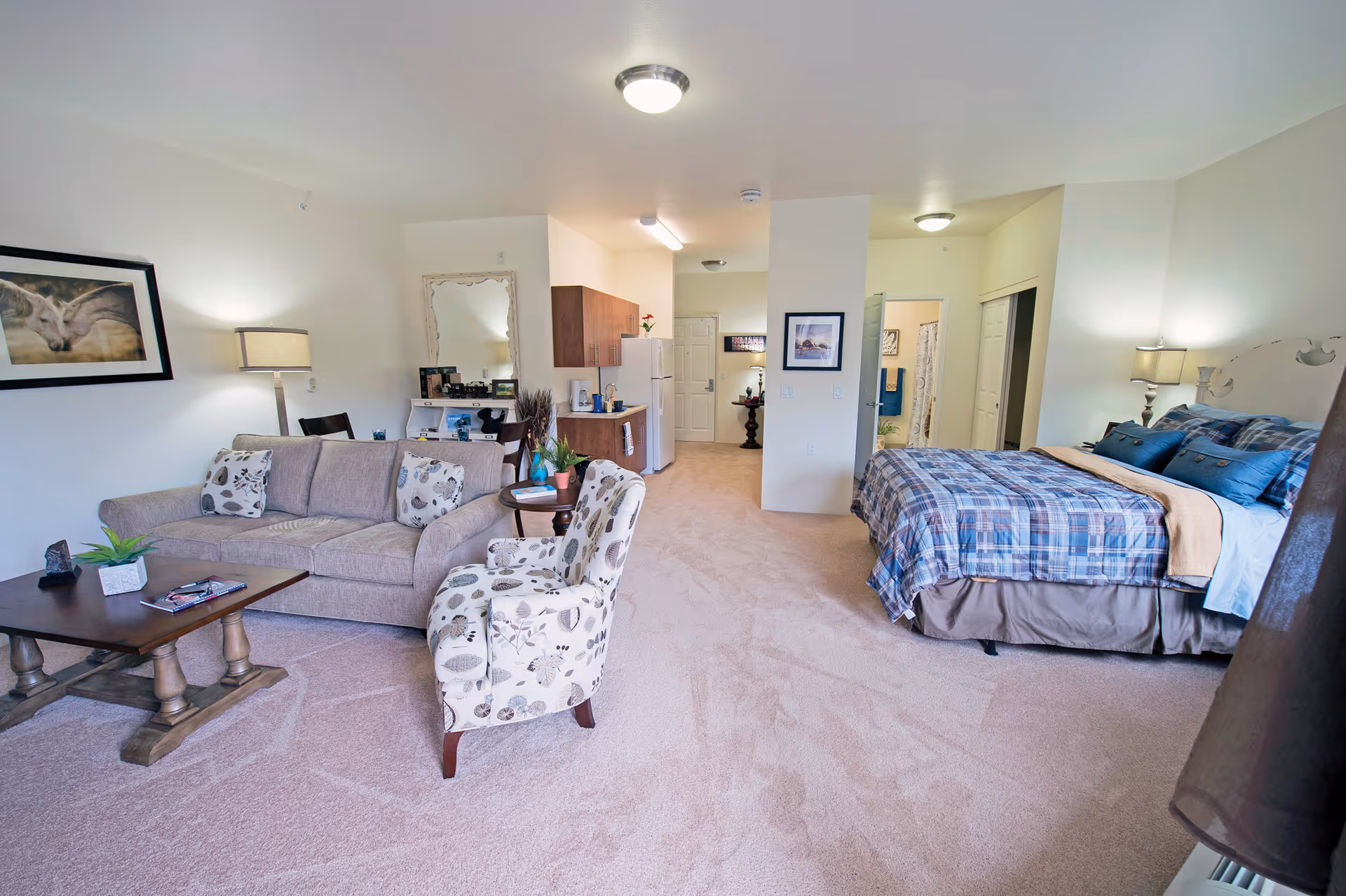A spacious studio apartment with a beige carpeted floor featuring a living area with a beige sofa, patterned armchair, and wooden coffee table. The room also includes a bed with blue plaid bedding, two bedside lamps, a small kitchen area with wooden cabinets, a refrigerator, and a coffee maker. A large mirror and framed artwork decorate the walls.