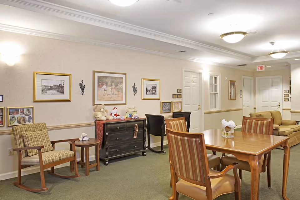 Well-lit communal living area with a dining table and chairs, armchairs and a sofa, framed artwork on the walls, and a dresser topped with stuffed animals.
