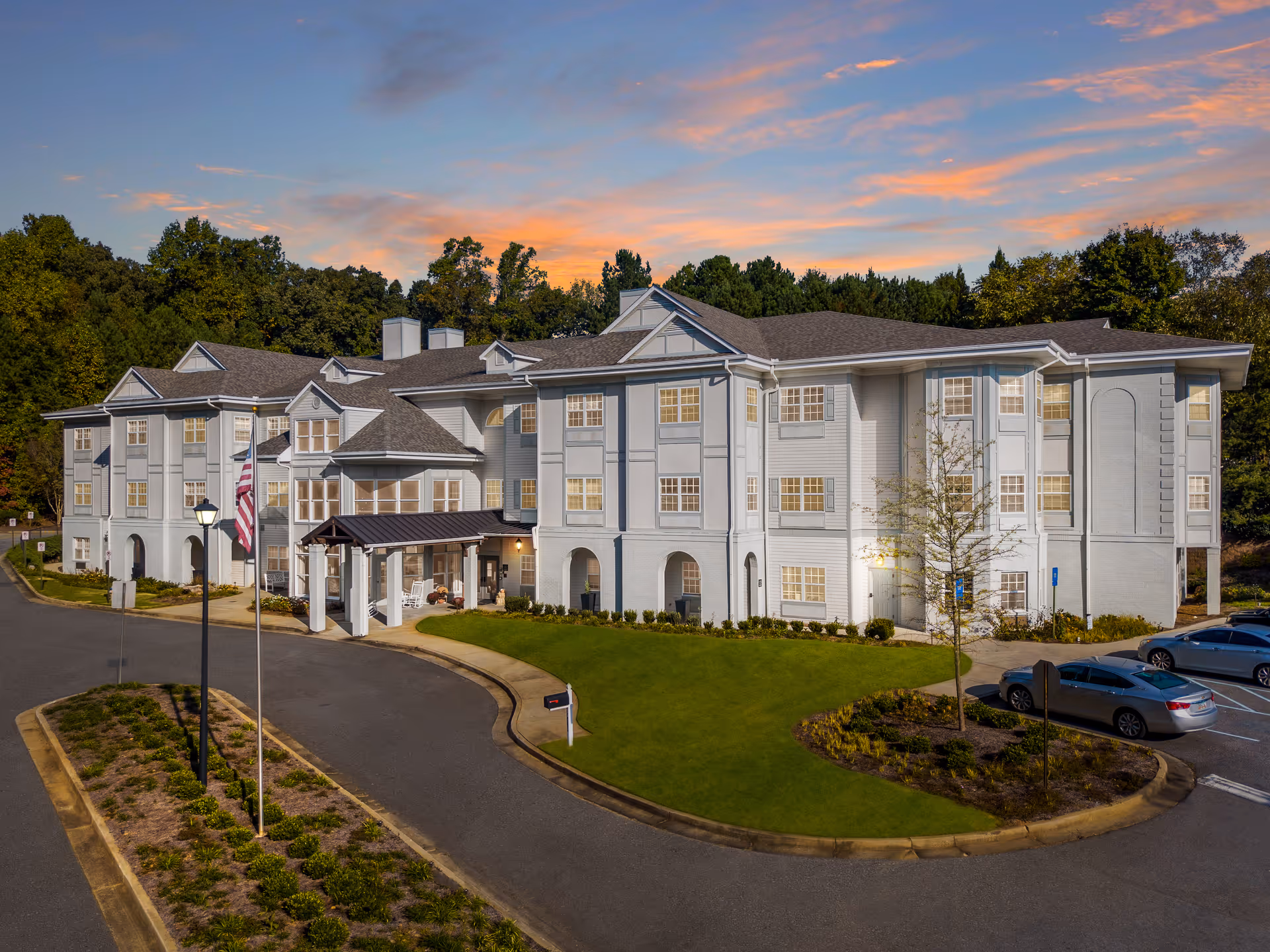 Exterior view of a large, three-story senior living facility building at sunset with a well-maintained lawn, a driveway, parked cars, and an American flag near the entrance.
