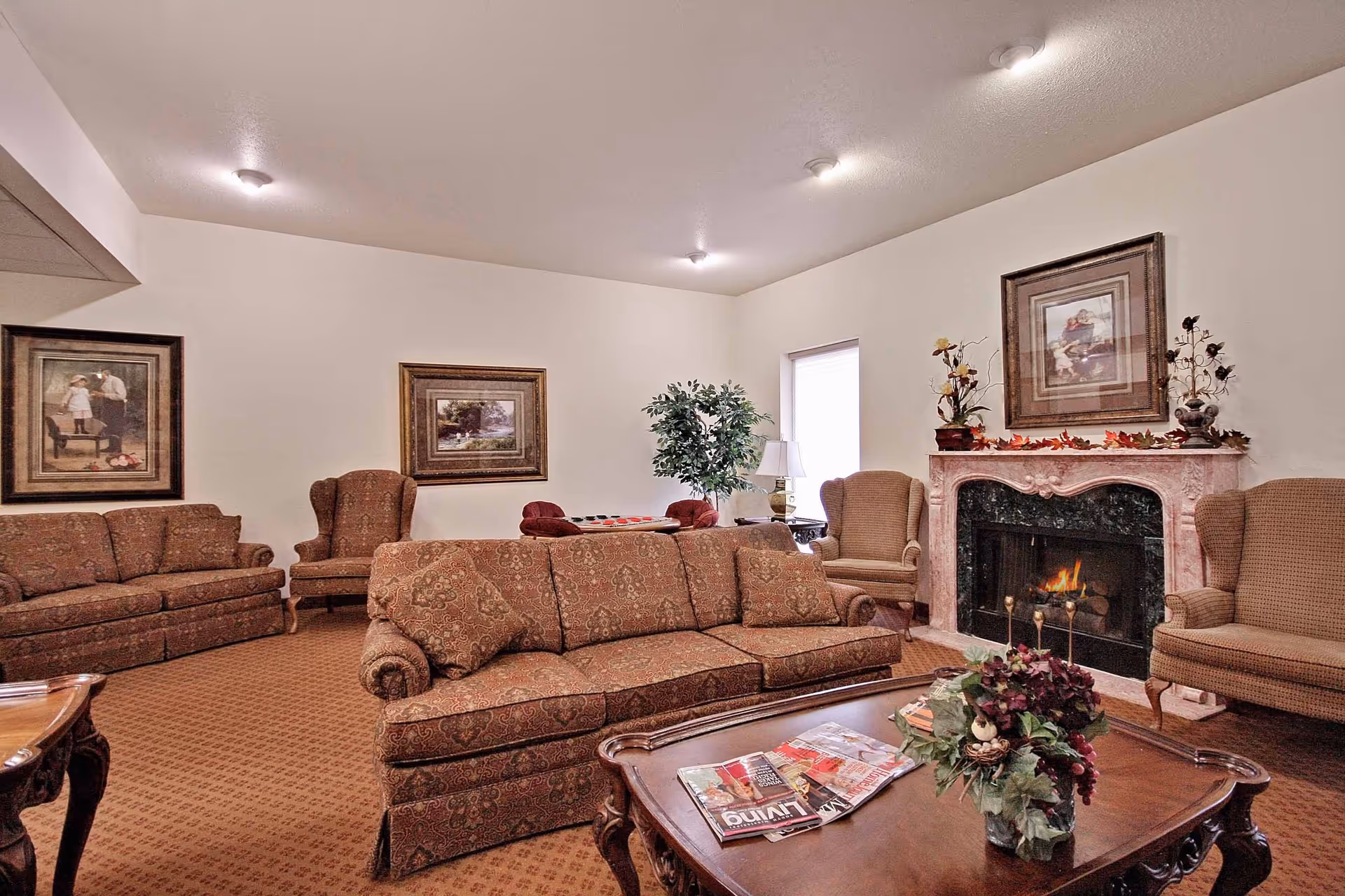 A cozy living room with patterned sofas and armchairs arranged around a wooden coffee table with magazines and a floral centerpiece. There is a lit fireplace with decorative items on the mantel, framed artwork on the walls, and a potted plant near a window.