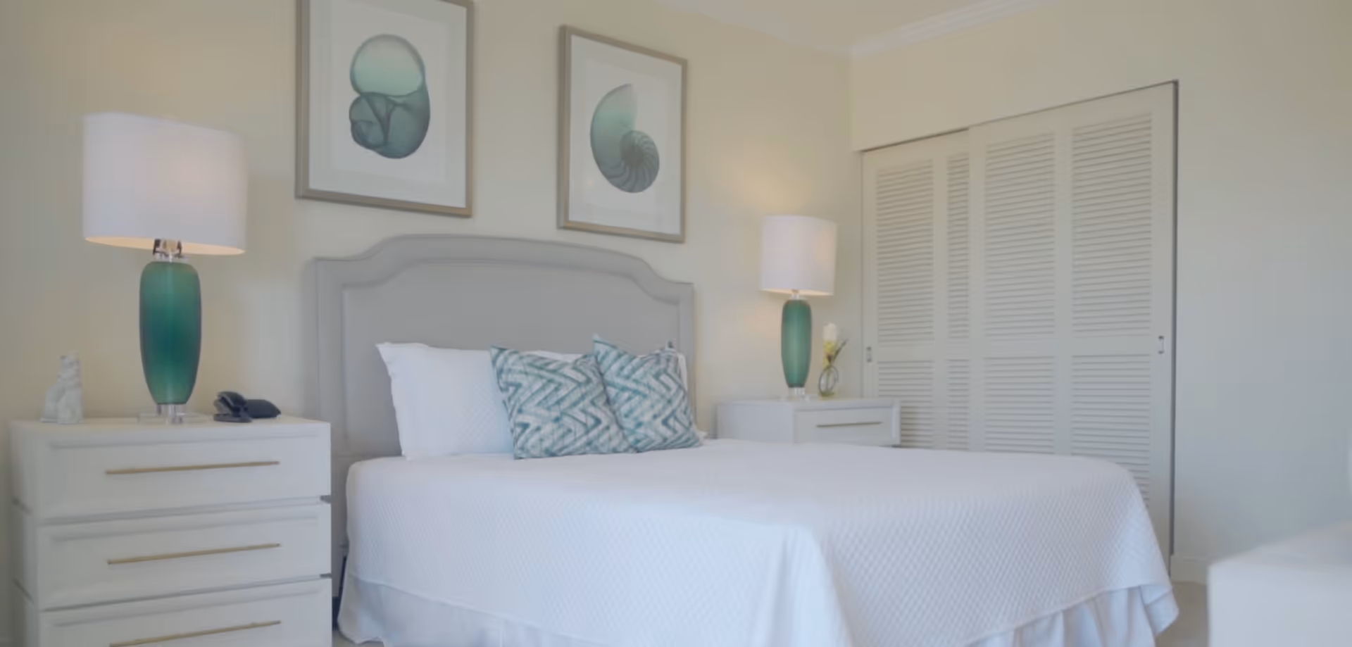 A neatly made bed with a light gray upholstered headboard, white bedding, and two patterned blue-green pillows. On each side of the bed are white nightstands with green glass lamps and decorative items. Above the bed are two framed abstract art pieces. A white closet with sliding louvered doors is visible in the background.