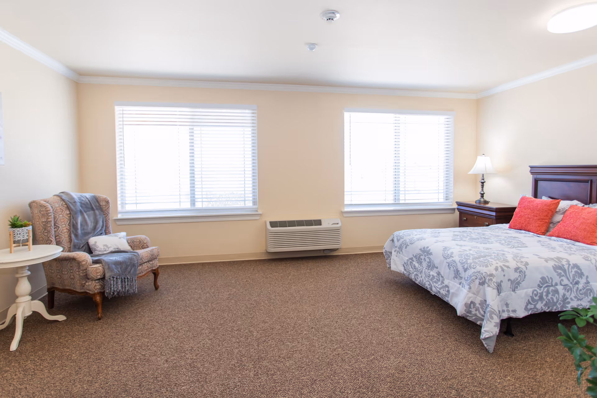 A bright and spacious bedroom with two large windows covered with white blinds. The room features a patterned armchair with a gray throw blanket and a decorative pillow, a small round white side table with a small plant, and a bed with a white and gray patterned comforter and three red pillows. There is a wooden nightstand with a lamp beside the bed, and the floor is covered with brown carpet.