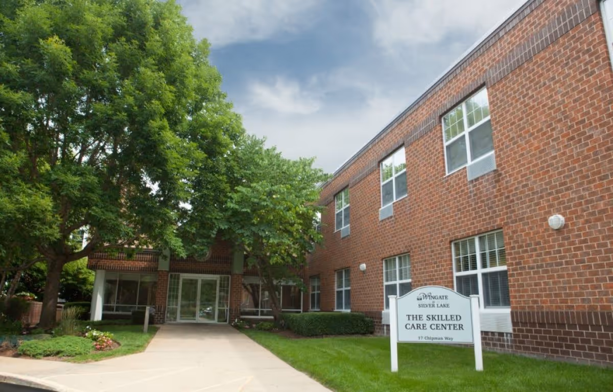 Exterior view of Wingate at Silver Lake Skilled Care Center showing a brick building with multiple windows, a concrete walkway leading to glass entrance doors, and green trees and landscaping around the entrance.