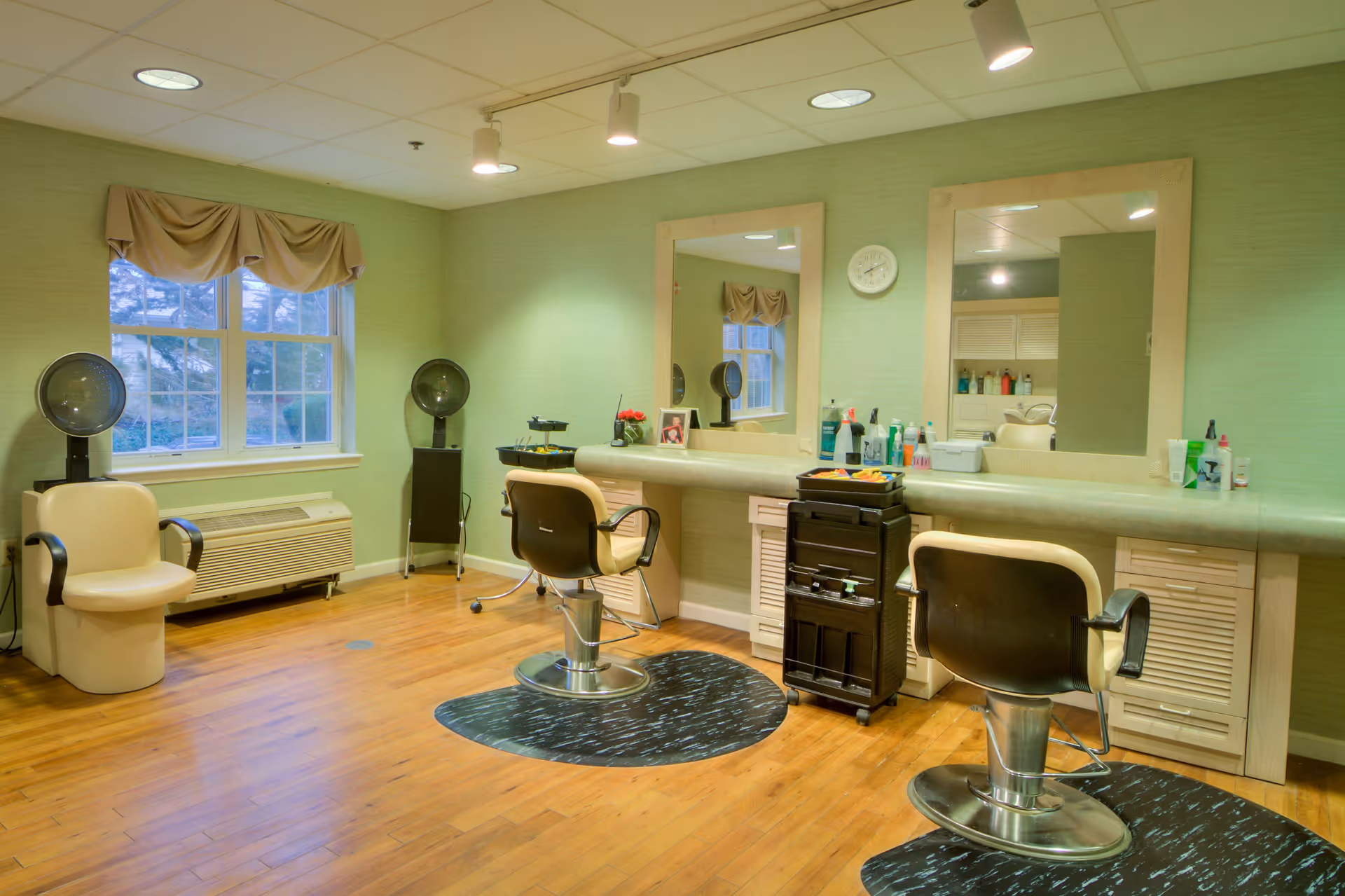 Interior view of a hair salon area with two salon chairs in front of a long counter with large mirrors. The room has light green walls, wooden flooring, and two hair dryers near a window with a beige valance. Various hair care products and tools are placed on the counter and a rolling cart.