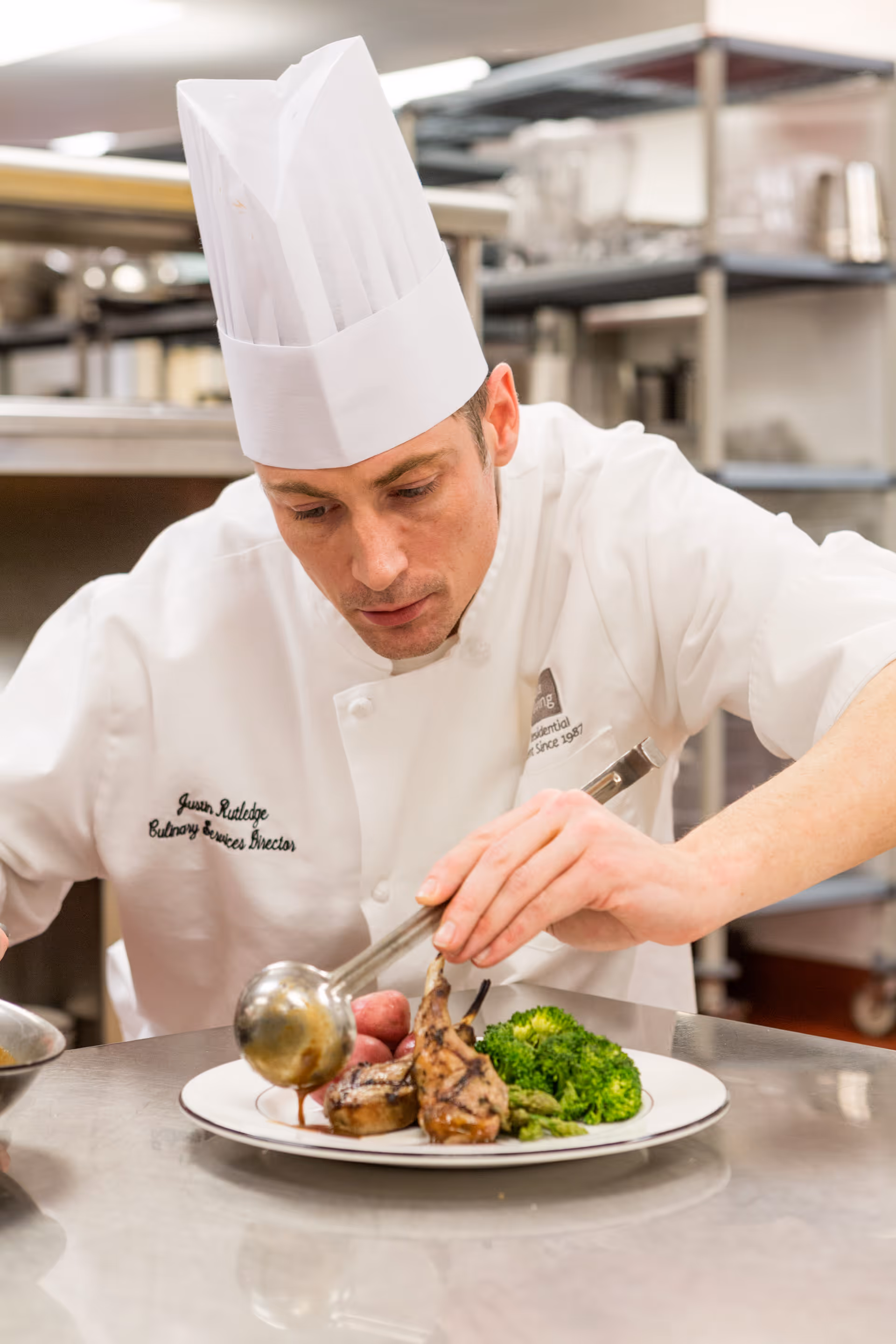 A chef wearing a white uniform and tall white chef hat carefully pours sauce onto a plated meal consisting of grilled meat, broccoli, asparagus, and red potatoes in a professional kitchen setting.