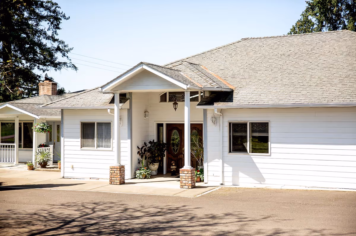 Front exterior view of a single-story white building with a gabled roof, a covered entrance supported by two white columns with brick bases, potted plants near the entrance, and windows with blinds. There are trees and a clear sky in the background.