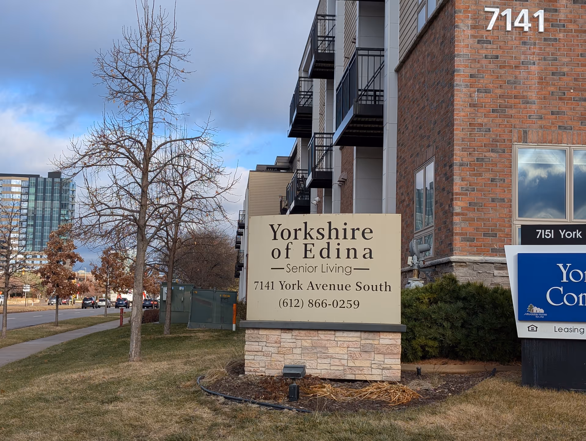Outdoor view of Yorkshire of Edina Senior Living facility sign with address 7141 York Avenue South and phone number (612) 866-0259, next to a multi-story building with balconies and a tree without leaves on a grassy area.