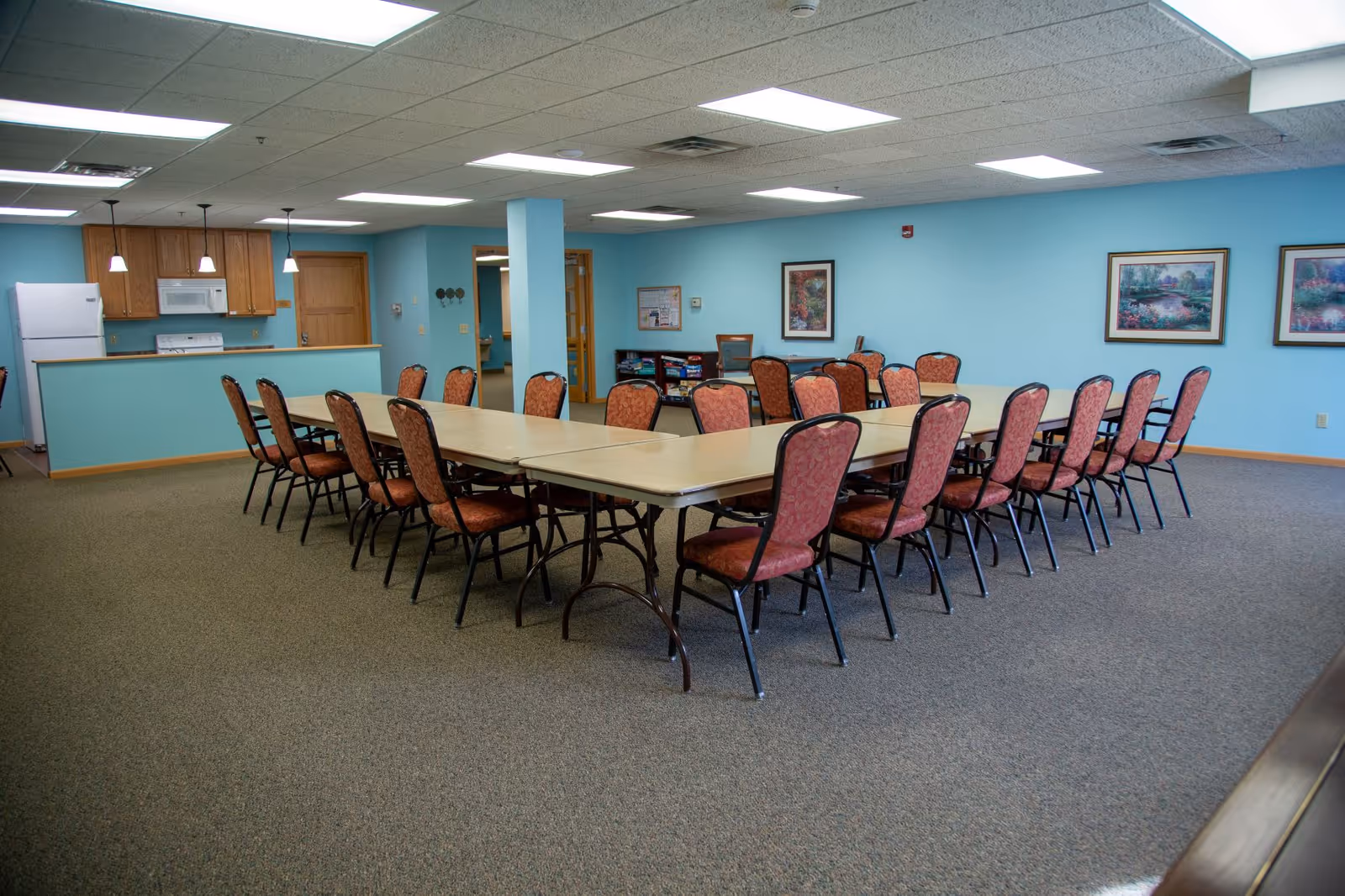 A spacious room with a large rectangular table surrounded by multiple red cushioned chairs. The walls are painted light blue and decorated with framed artwork. In the background, there is a small kitchen area with a refrigerator, microwave, stove, and wooden cabinets. The room is well-lit with ceiling lights and has carpeted flooring.