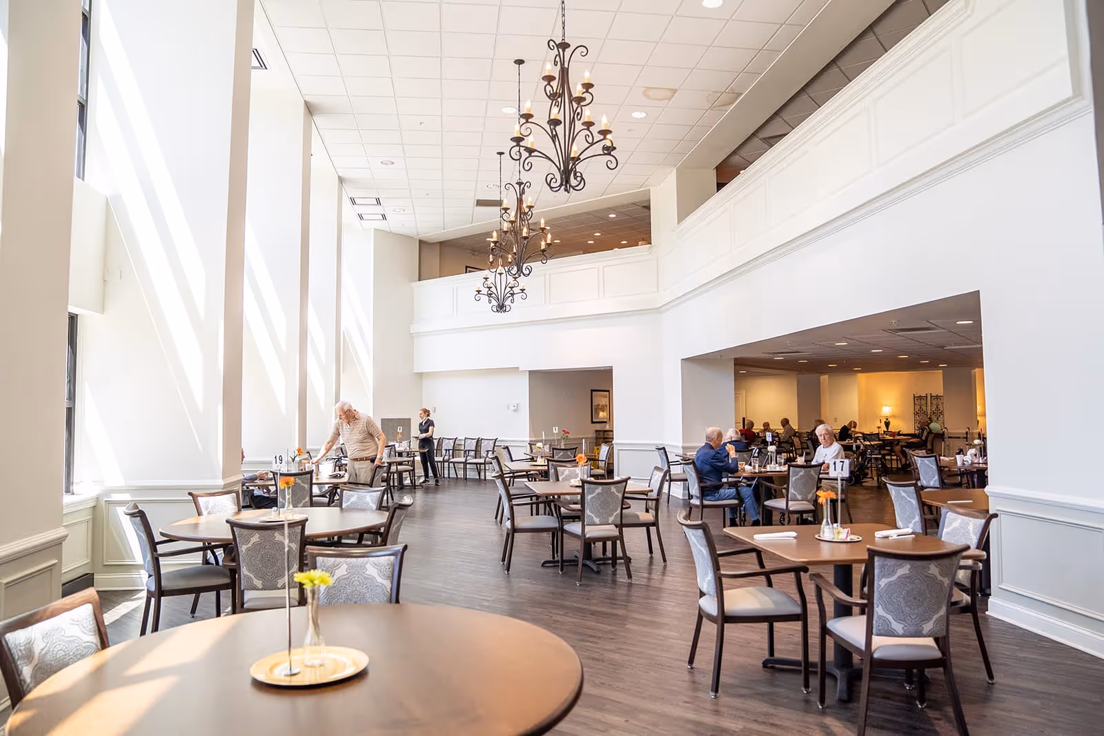 Bright and spacious dining room with large windows letting in natural light, several round tables with chairs, a few elderly people seated and dining, and elegant chandeliers hanging from the high ceiling.