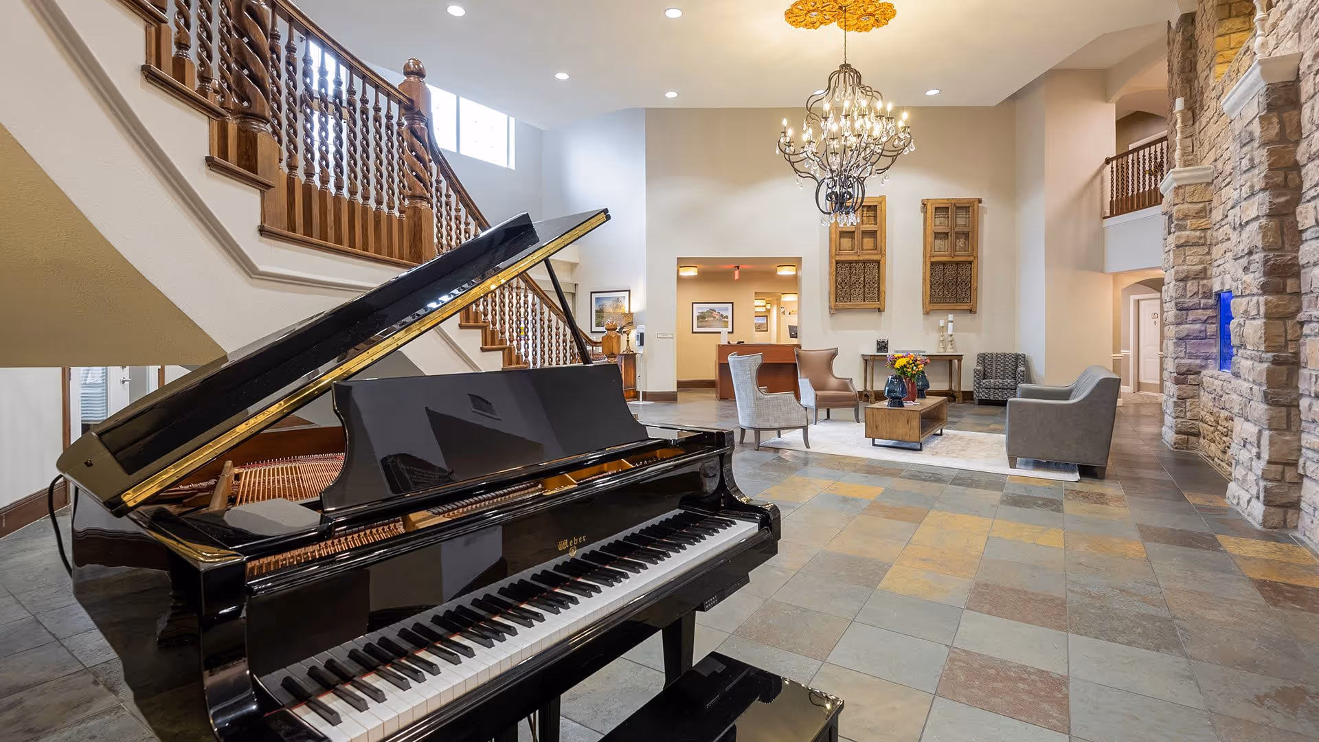 Spacious and elegant living room area with a grand black piano in the foreground, a wooden staircase with ornate railings, a chandelier hanging from the ceiling, and comfortable seating arranged around a coffee table with flowers. The room features stone walls, large tiled flooring, and warm lighting.