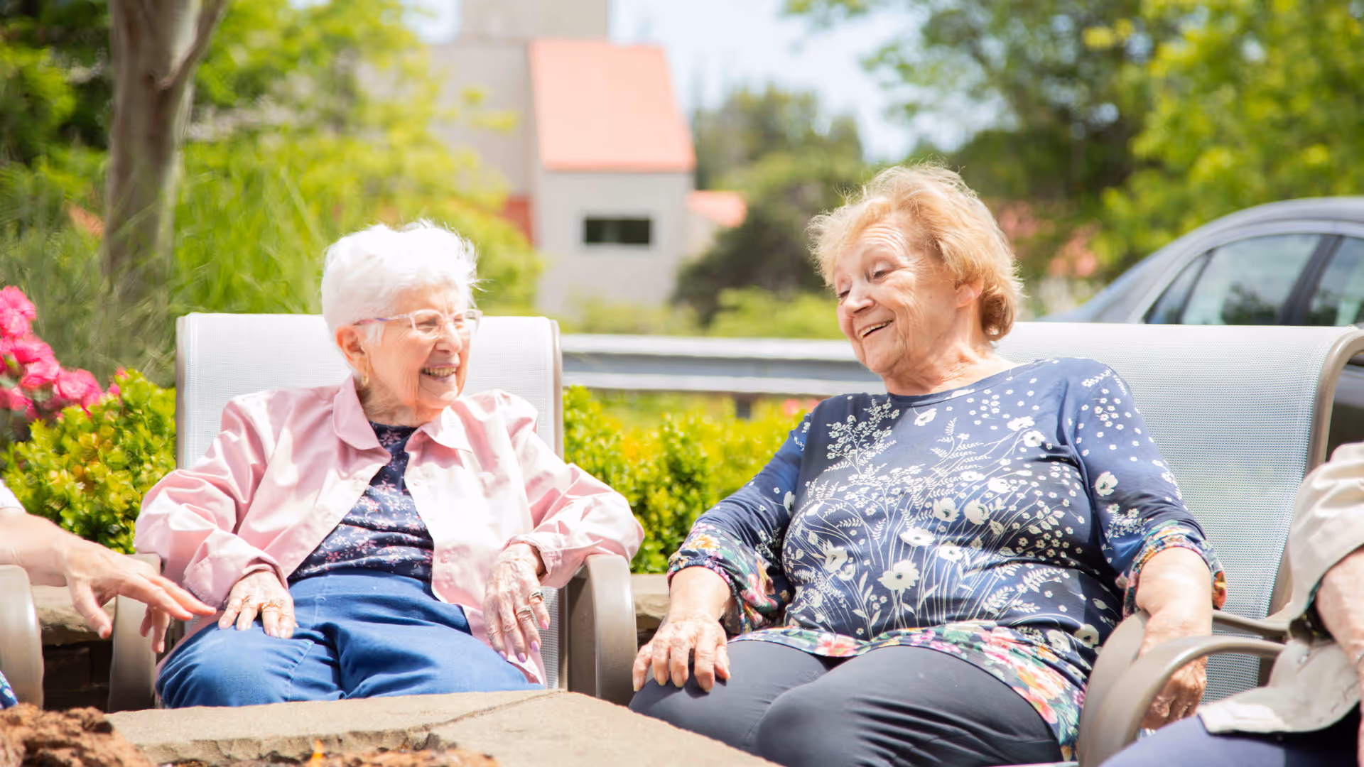 Two elderly women sitting outdoors on patio chairs, smiling and engaging in conversation, surrounded by greenery and flowers with a building and car visible in the background.