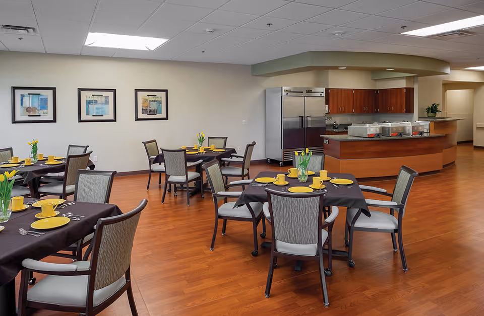 A dining room in a senior living facility with several round tables covered with dark tablecloths, each set with yellow plates, cups, and silverware. There are chairs around the tables and vases with yellow flowers as centerpieces. In the background, there is a kitchen area with stainless steel refrigerators, wooden cabinets, and a counter with food warmers. The walls are decorated with framed artwork.