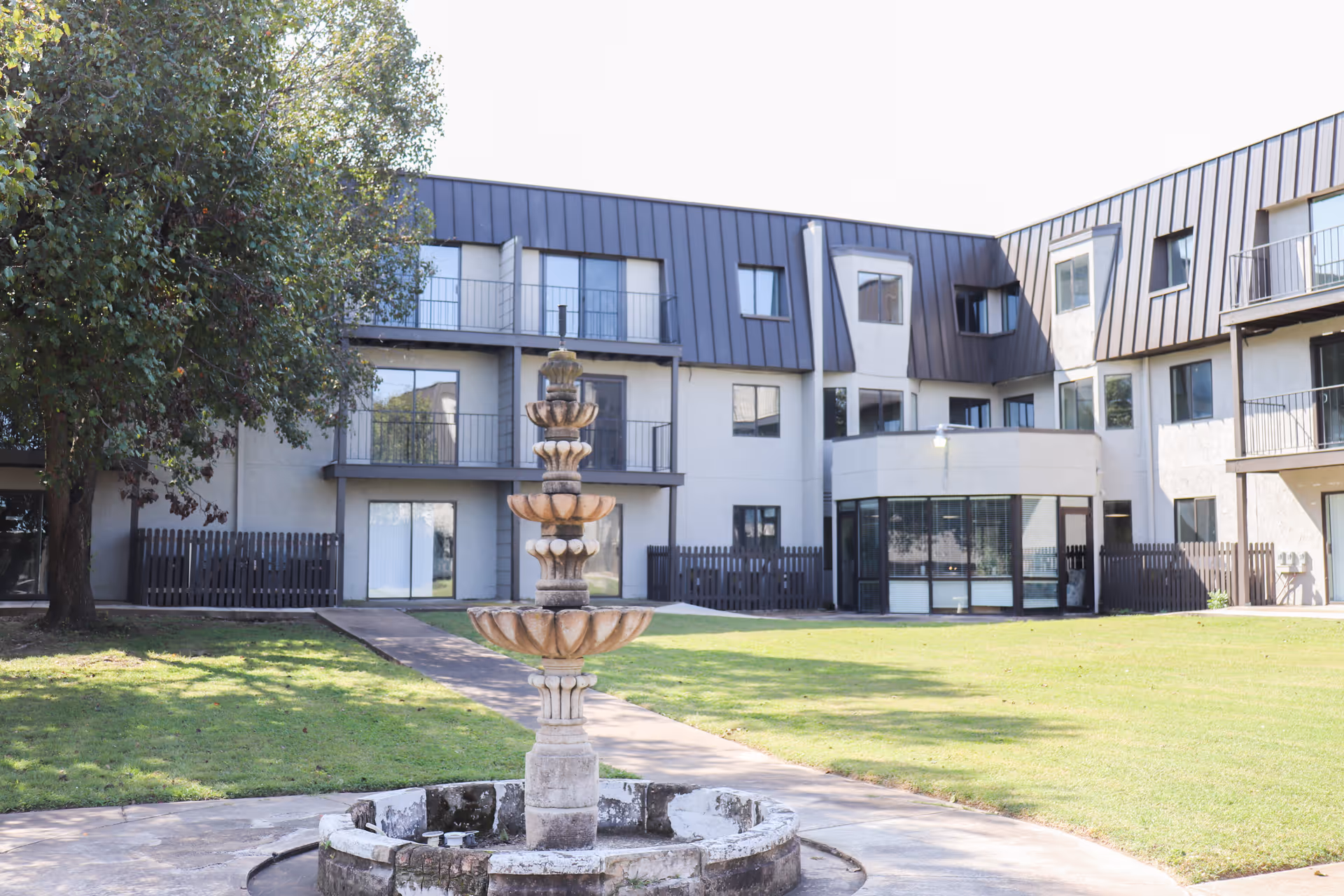 Outdoor courtyard area of a multi-story retirement community building with a central stone fountain, green lawn, a large tree on the left, and balconies on the upper floors.