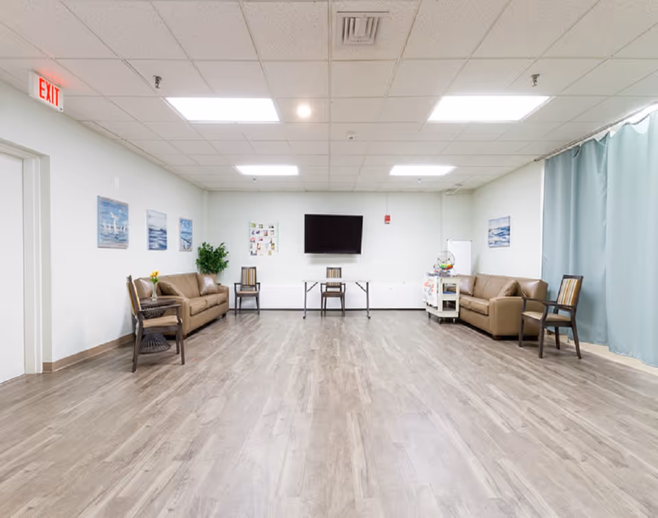A spacious senior living lounge with sofas, chairs, a wall TV, and a blue curtain.