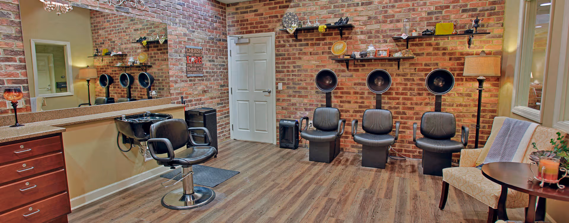 Interior of a hair salon area with brick walls and wooden flooring. There are three black hair drying chairs with hooded dryers against the wall, a single salon chair in front of a counter with a sink, and a cozy seating area with a patterned armchair, a small round table with a candle, and a floor lamp. Shelves on the wall hold decorative items and hair products.