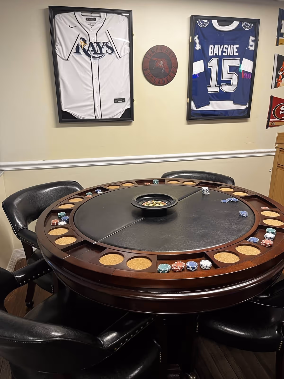 Round poker table with chips and chairs in a room decorated with framed sports jerseys on the wall.