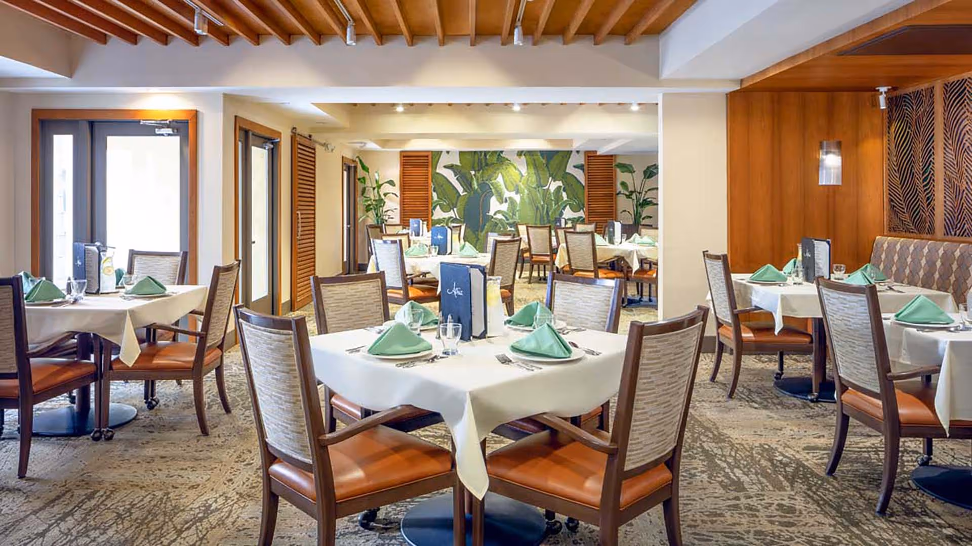 A well-lit dining room with multiple tables covered in white tablecloths, each set with green folded napkins, glassware, and menus. The room features wooden chairs with cushioned seats, a patterned carpet, and a wall decorated with large green leaf artwork and wooden panels.