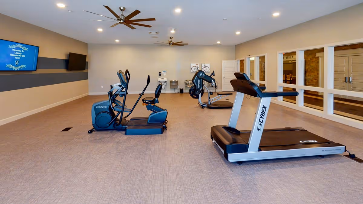 Interior fitness room with treadmills, ellipticals and stationary bikes on a carpeted floor with ceiling fans.