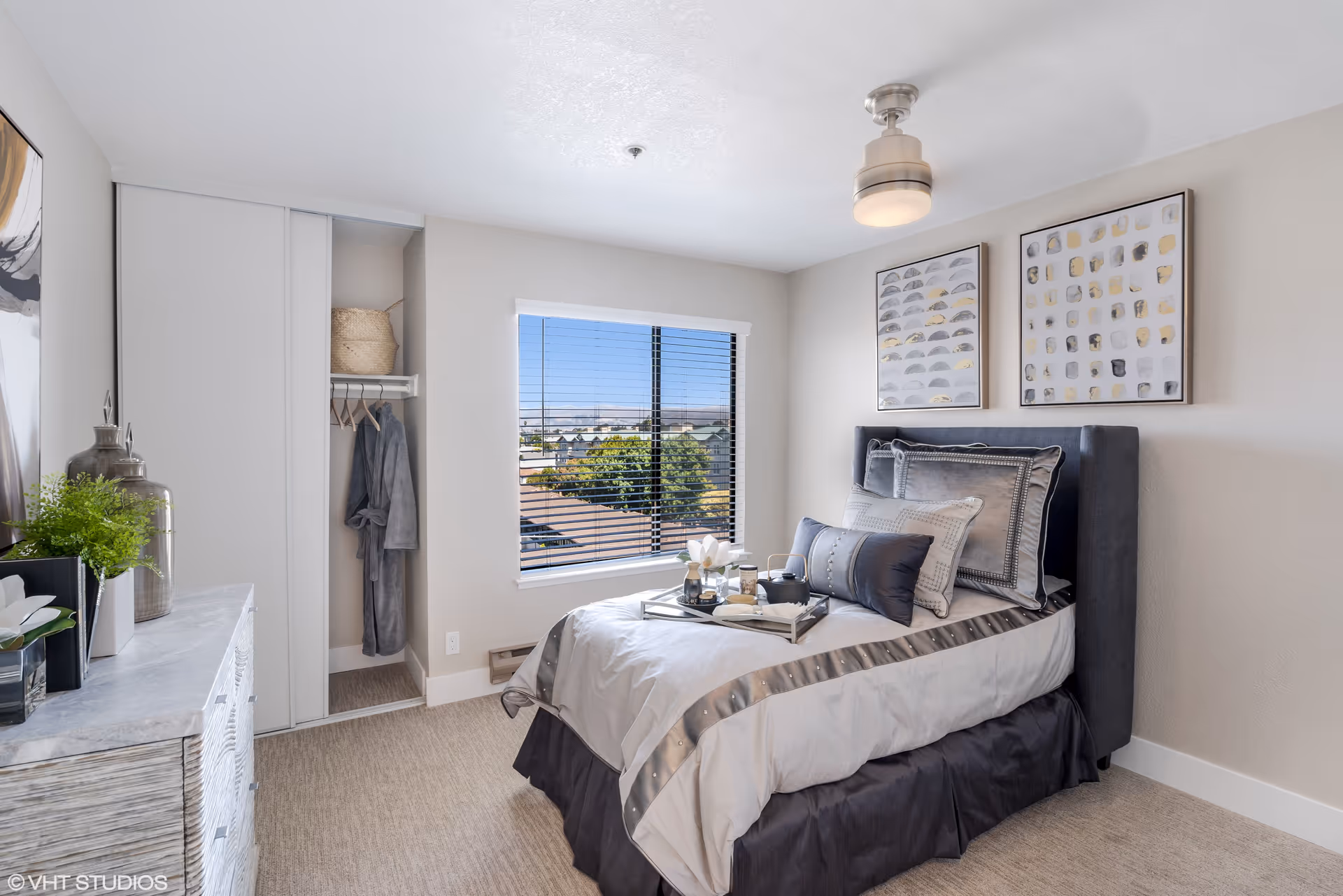A bright and neatly arranged bedroom with a single bed dressed in white and gray bedding, decorative pillows, and a tray with tea set on top. The room features a window with blinds showing a view of trees and rooftops outside. There is a small open closet with a hanging robe and a woven basket on the shelf. A dresser with decorative items and plants is positioned on the left side, and two framed abstract artworks hang above the bed. The ceiling has a modern light fixture.