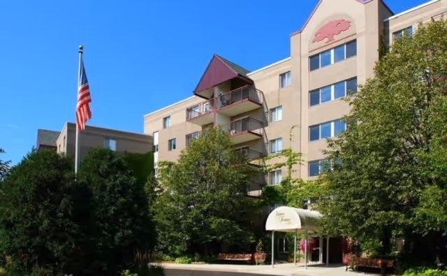 Exterior view of Vernon Terrace of Edina, a multi-story senior living facility building with balconies, surrounded by trees and greenery, an American flag on a flagpole, and a covered entrance.