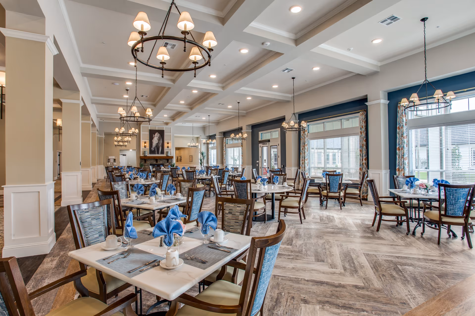 A spacious and well-lit dining room in a senior living facility with multiple tables set with blue napkins, white cups, and silverware. The room features large windows with floral curtains, elegant chandeliers hanging from a coffered ceiling, and a fireplace with a horse painting above it in the background.