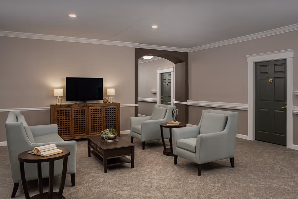 A cozy living room area in an assisted living facility featuring a flat-screen TV on a wooden cabinet with two table lamps on either side. The room has three light blue armchairs and one matching loveseat arranged around a dark wooden coffee table and two small side tables. The walls are painted beige with white trim, and there are two dark green doors visible in the background.