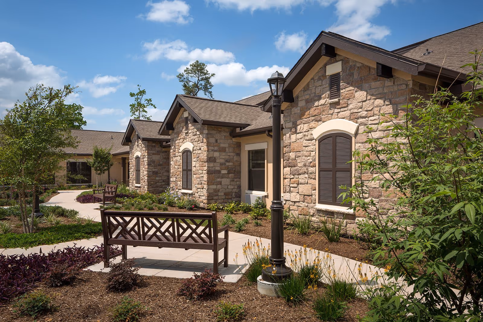 Outdoor courtyard area of a senior living facility with stone exterior walls, a paved walkway, benches, a lamppost, and landscaped plants under a blue sky with scattered clouds.