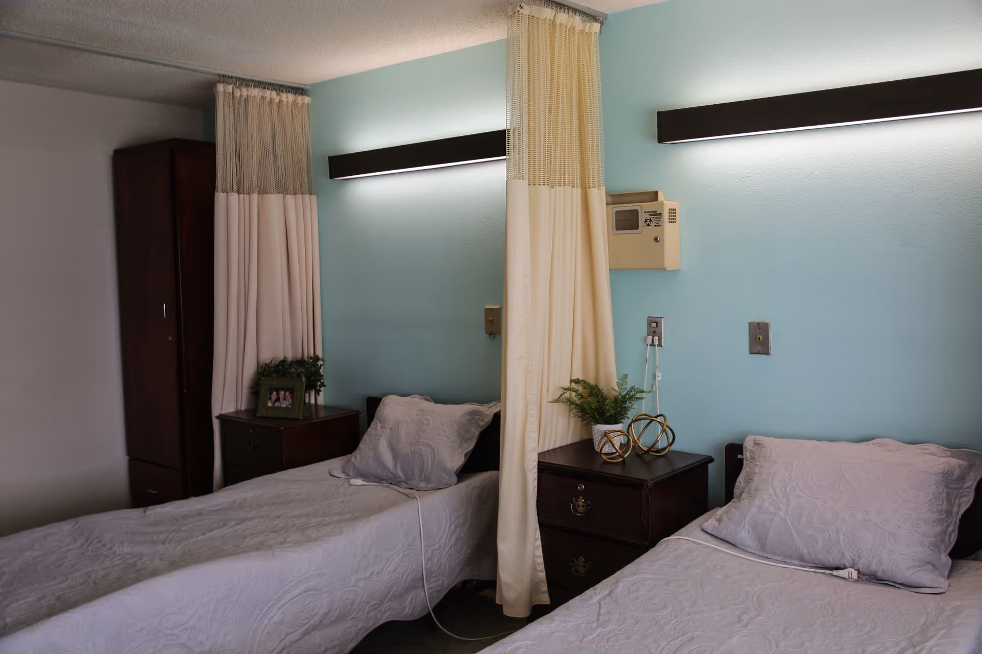 A room with two single beds separated by a privacy curtain. Each bed has a light purple bedspread and pillow. Between the beds is a dark wooden nightstand with decorative items and a small plant. The walls are painted light blue, and there are long horizontal lights above each bed. A tall dark wooden cabinet is visible in the corner.