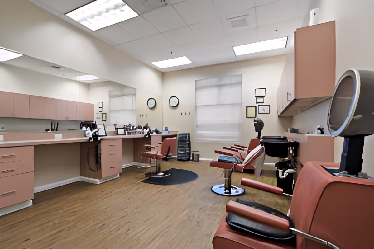 Interior of a salon room with multiple red salon chairs, hair washing stations, large mirrors on the walls, and pink cabinetry. The room has wooden flooring and bright overhead lighting.
