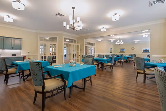 A spacious dining room with multiple tables covered in teal tablecloths, each set with glasses and napkins. The room has wooden flooring, light-colored walls, and several ceiling light fixtures. Chairs with patterned upholstery surround the tables, and there are glass-paneled double doors in the background.