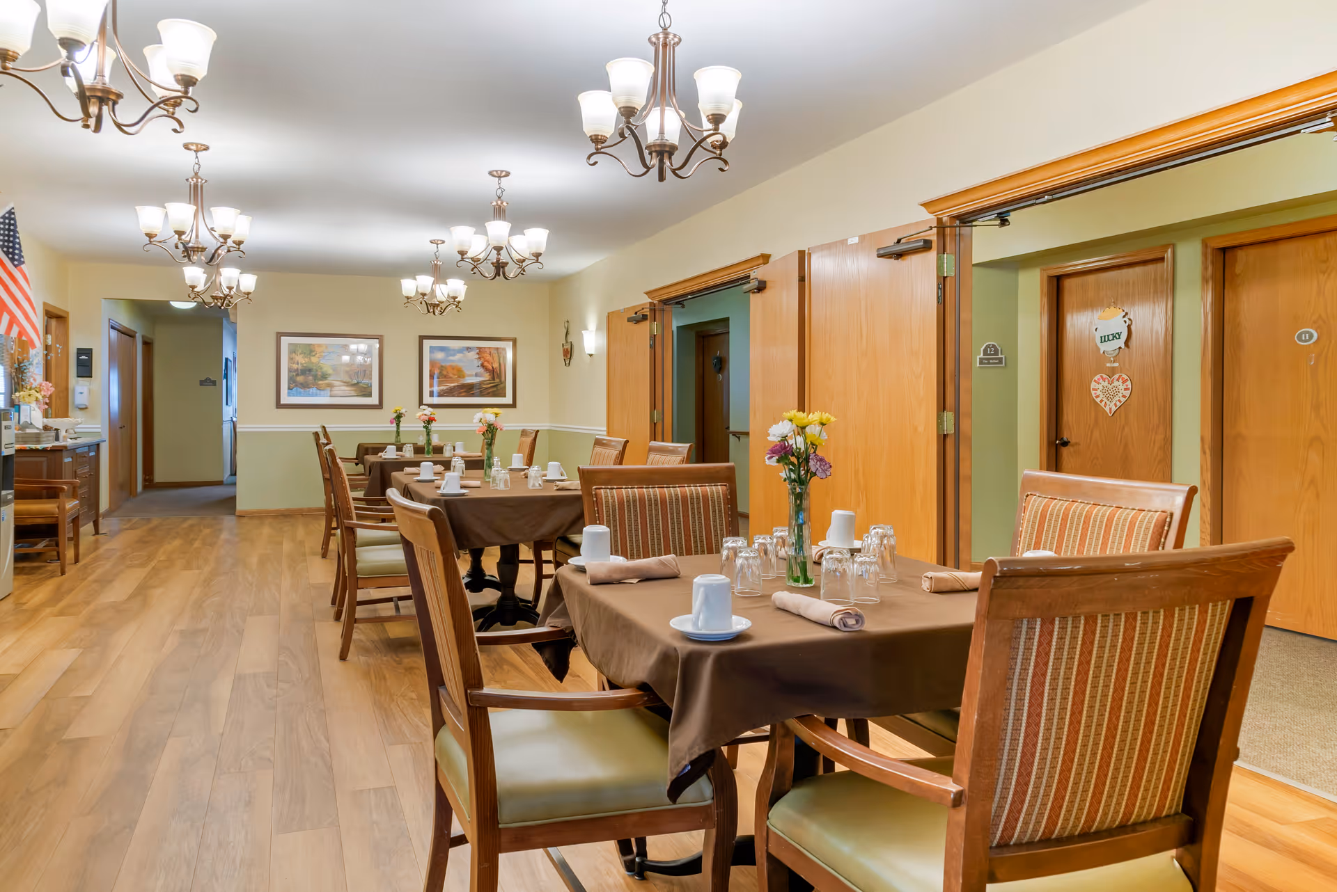 A dining room in a senior living facility with several tables covered with brown tablecloths, each set with cups, glasses, and napkins. Wooden chairs with striped cushions surround the tables. The room has wooden floors, multiple chandeliers, framed landscape paintings on the walls, and several wooden doors leading to other rooms.