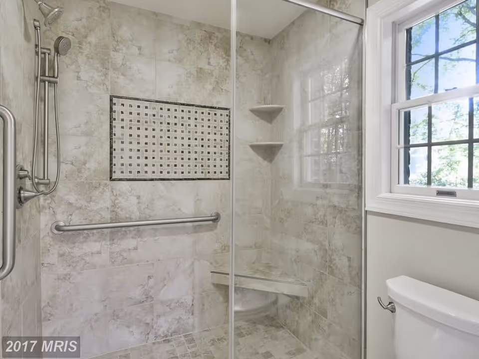 A modern bathroom shower area with beige marble tiles, a built-in corner bench, two corner shelves, a stainless steel grab bar, and a handheld showerhead. There is a window with white trim to the right and a white toilet partially visible.