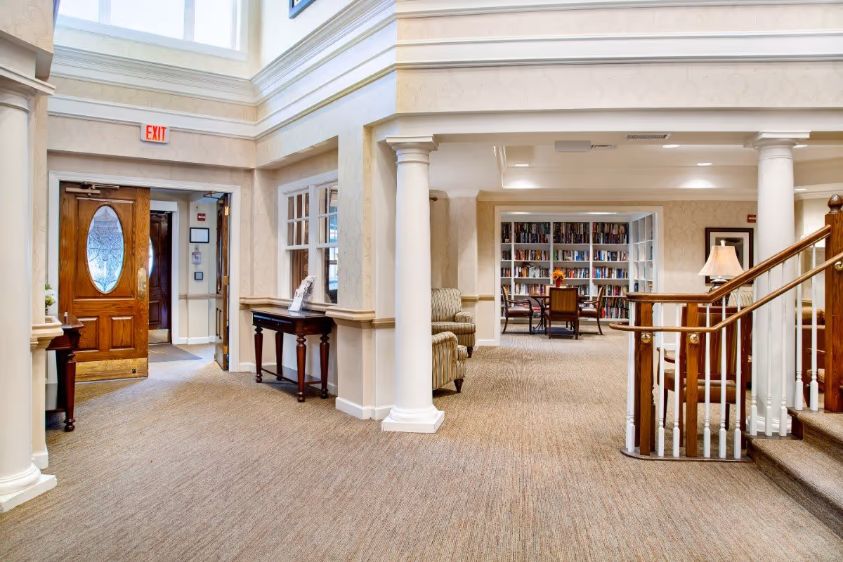 Interior view of a senior living facility lobby with columns, seating, a staircase and a book-lined reading area.