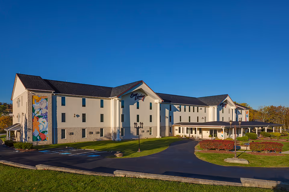Exterior front view of a three-story senior living building with a covered entrance, landscaped grounds, and a colorful mural on one side under a clear blue sky.