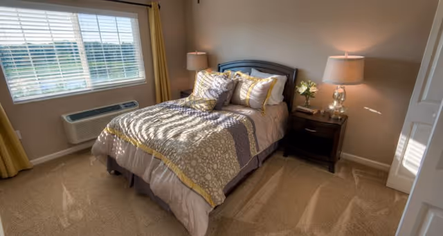 Sunlit bedroom with a neatly made bed, two bedside tables with lamps, a window with blinds and yellow curtains, and beige carpeting.