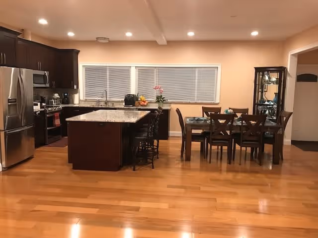 A spacious kitchen and dining area with wooden floors and beige walls. The kitchen features dark wood cabinets, stainless steel appliances including a refrigerator and stove, and a large island with a granite countertop and bar stools. The dining area has a glass-top table with six chairs and a glass-front cabinet displaying dishes. A window with closed blinds is above the kitchen sink.