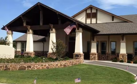 Exterior view of a single-story assisted living facility building with a covered entrance supported by large pillars. The building features stone accents along the lower walls and beige siding above. Several American flags are displayed on the building and in the surrounding landscaped lawn area.