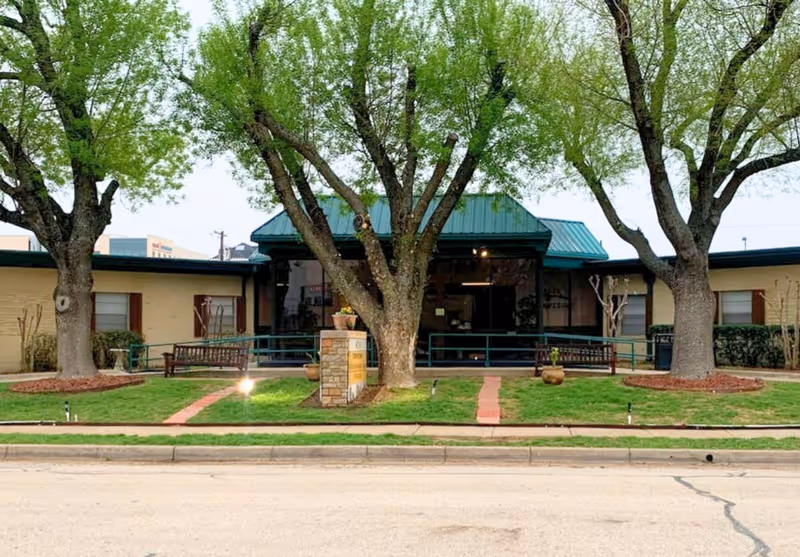 Front exterior of a single-story health and rehabilitation center with a green metal roof, large trees, benches, and a brick walkway to the entrance.