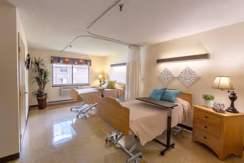 Shared patient room with two adjustable beds, bedside tables, lamps, a window, and a privacy curtain.