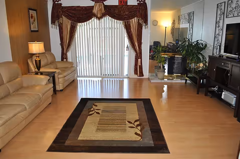 A living room with light wood flooring, a beige leather sofa and loveseat, a side table with a lamp, a patterned area rug in the center, large vertical blinds covering a sliding glass door with dark red curtains, several potted plants near a fireplace, and a dark wood entertainment center with a flat-screen TV on the right wall.