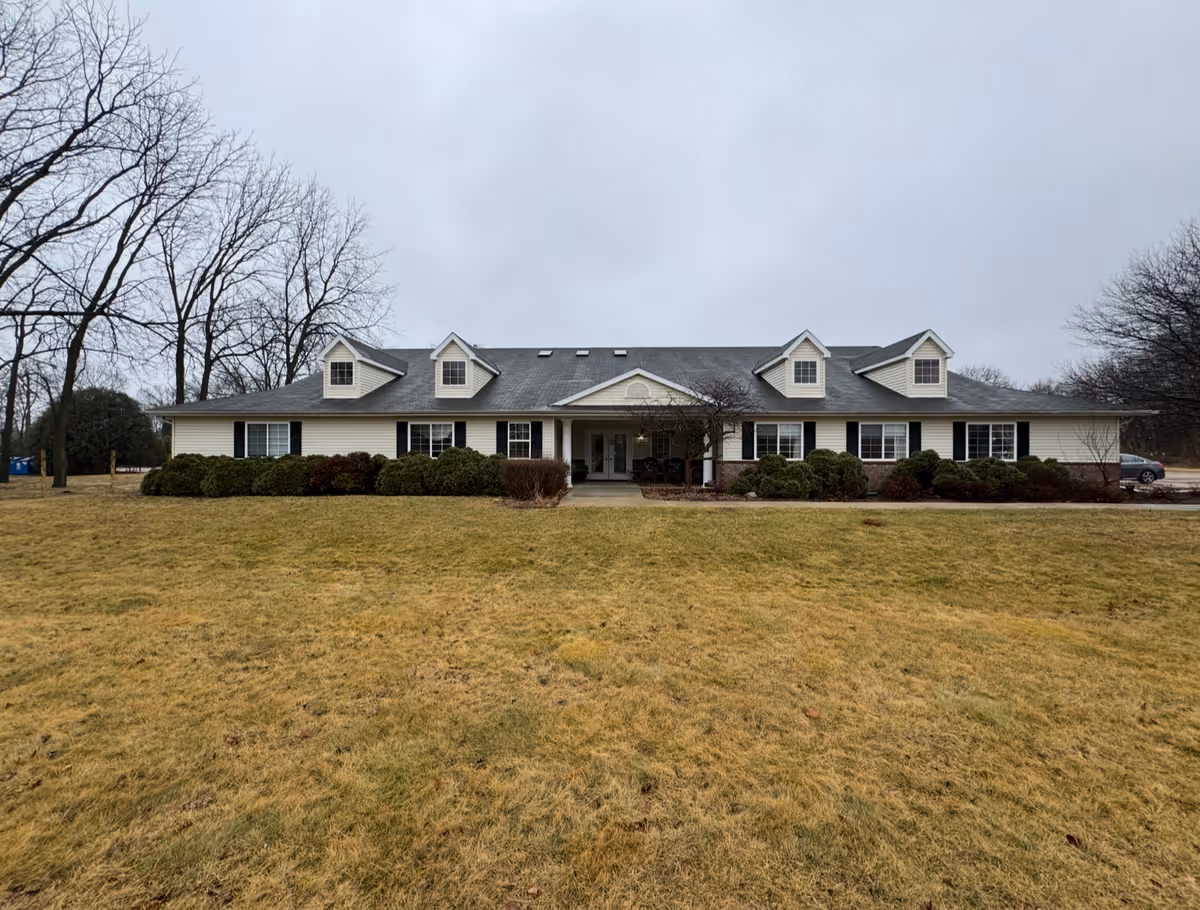 Front exterior view of a single-story senior living facility building with a gray roof, white siding, and several dormer windows. The building is surrounded by trimmed bushes and a large grassy lawn with leafless trees in the background under an overcast sky.
