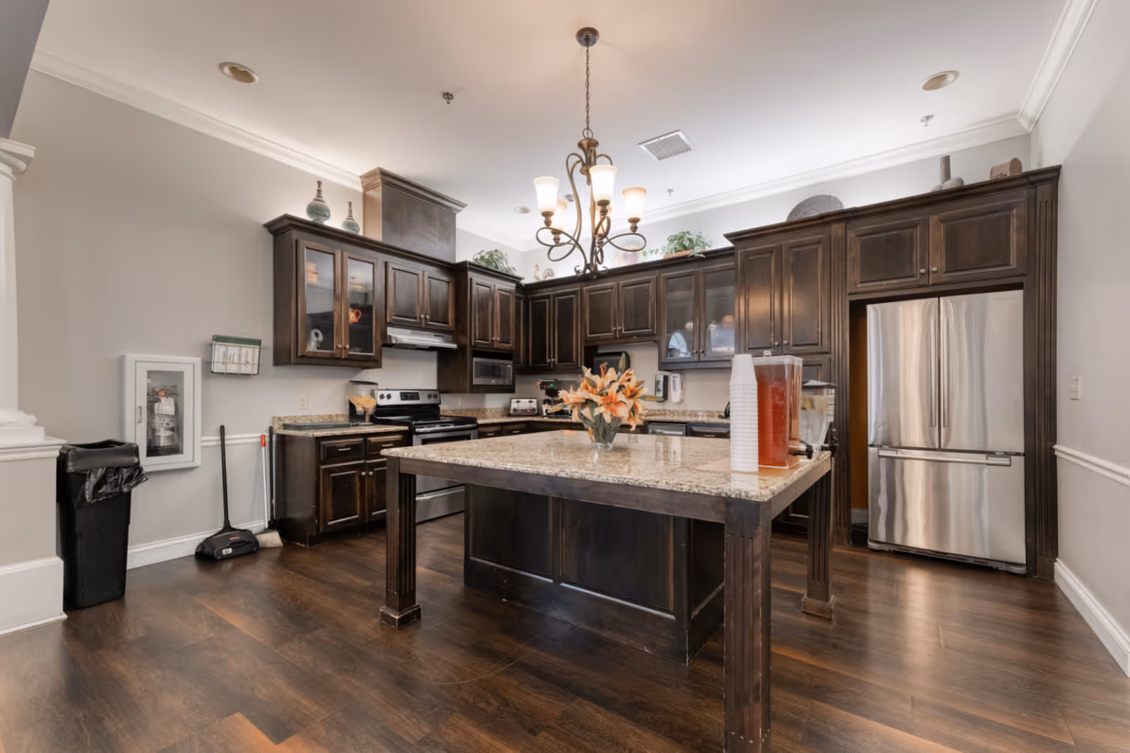 A spacious kitchen with dark wooden cabinets and a large central island topped with granite. The kitchen features stainless steel appliances including a refrigerator and stove. A chandelier hangs above the island, which has a vase of flowers and beverage dispensers with cups. The floor is dark wood, and the walls are painted light gray with white trim.
