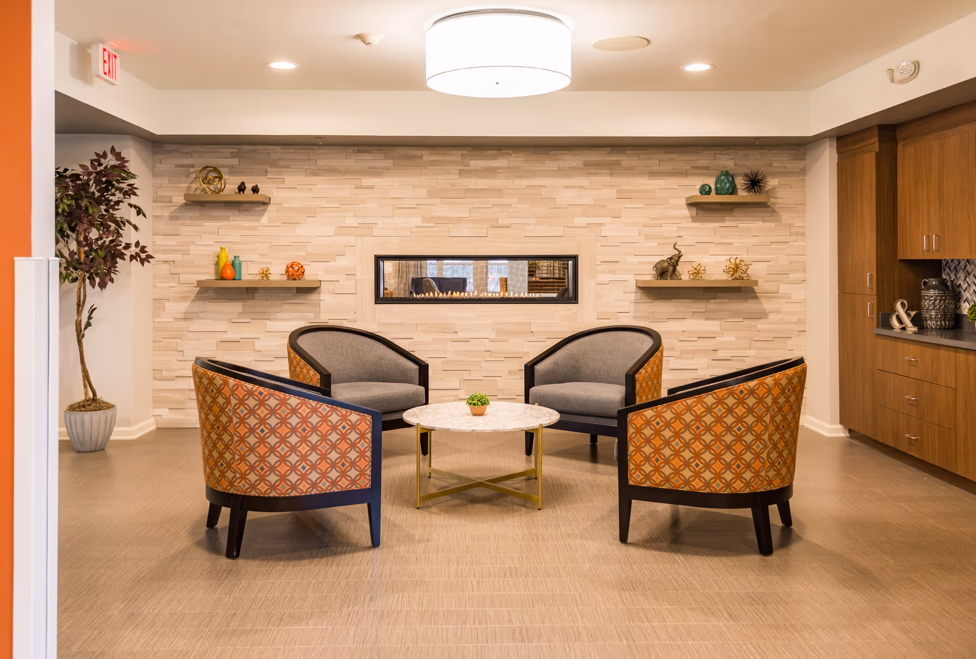 A cozy seating area with four patterned armchairs arranged around a round marble-top coffee table with a small plant. The background features a modern stone wall with a rectangular built-in fireplace and decorative shelves holding various ornaments. To the right, there are wooden cabinets and a countertop with decorative items. A potted plant is visible on the left side of the image.