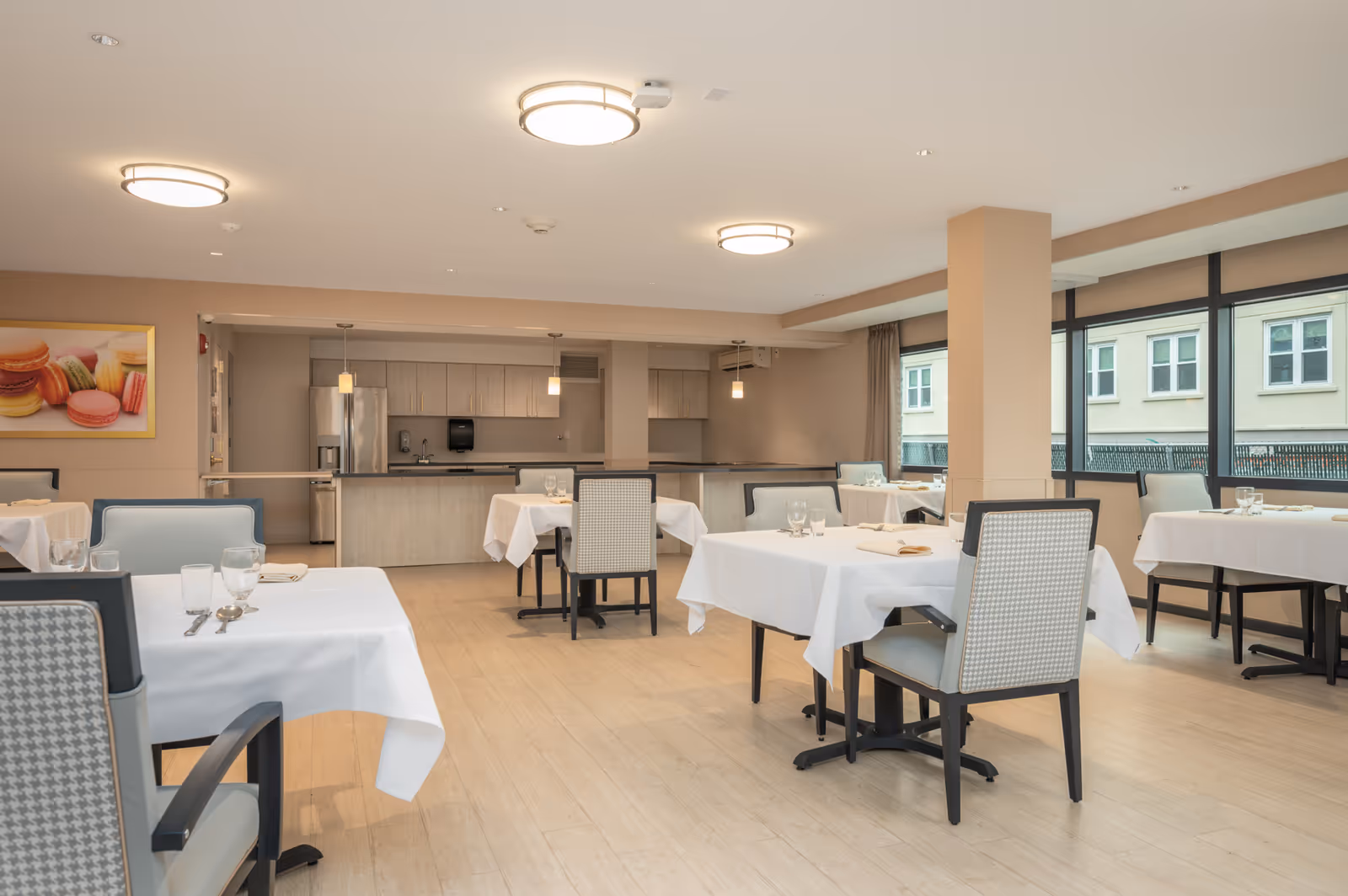 Spacious dining room with tables set with white tablecloths and chairs, a serving counter in the back, and large windows.