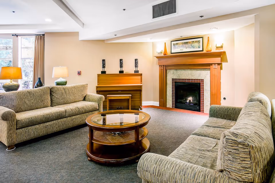 A cozy living room with two beige patterned sofas facing each other, a round wooden coffee table with a glass top in the center, a wooden piano against the wall with three decorative candle holders on top, and a fireplace with a wooden mantel and a framed painting above it. There are two table lamps on side tables next to the sofas and large windows with curtains letting in natural light.