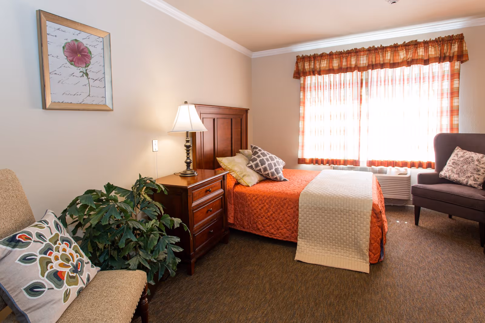 Cozy bedroom with a single bed dressed in an orange quilt, wooden nightstand and lamp, armchair, and a window with patterned curtains.